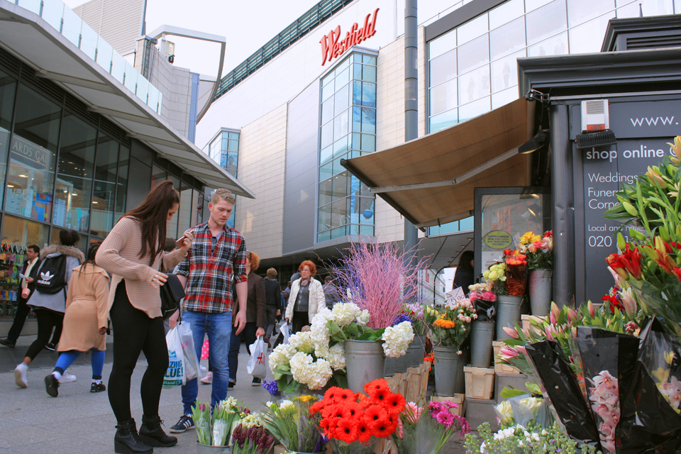 Two people looking at flower bouquets, outside the Westfield, Shepherd's Bush, a large building.